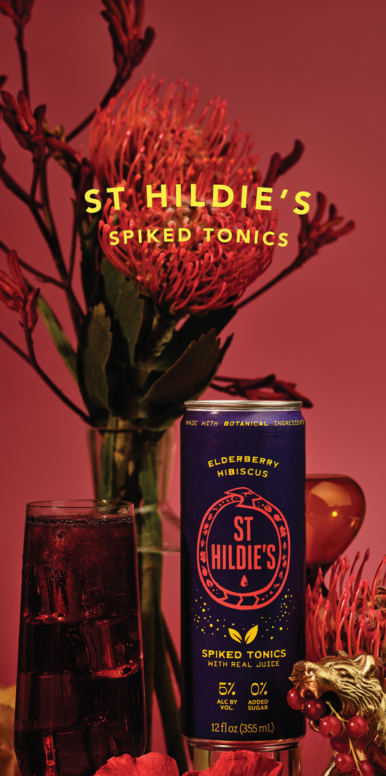 A dark purple can sits on a red background surrounded by red botanicals and flowers, and a glass of dark liquid and ice sits to the left of the can. 